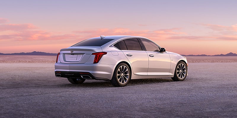 A stylish white Cadillac sedan is parked on an open desert road at sunset, highlighting its sleek rear design, sharp taillights, and dual exhaust tips.