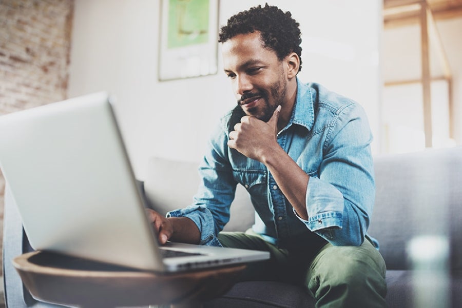 A man with a thoughtful expression sits on a couch, wearing a denim shirt and green pants, as he looks at a laptop screen in a well-lit, modern living space.